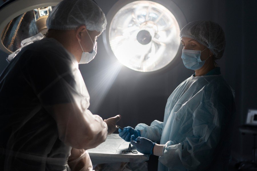 Two medical professionals in surgical attire prepare instruments in an operating room under bright surgical lights.