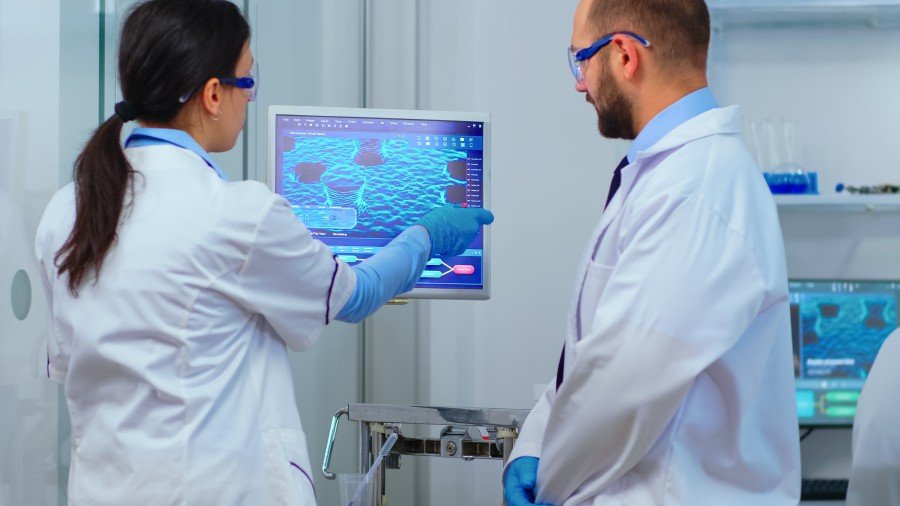 Two laboratory scientists in protective coats and gloves examining a computer display showing a microscopic biological image.