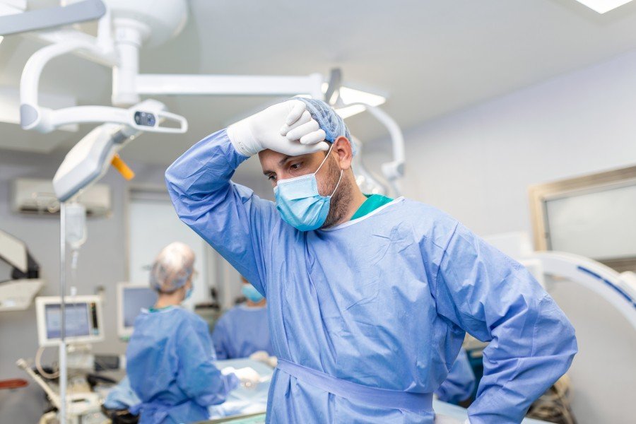 Robotic Surgeon in surgical scrubs and mask standing in an operating room, appearing fatigued.