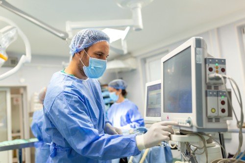 Robotic Surgeon monitoring medical equipment in an operating room while wearing blue surgical scrubs, gloves, a mask, and a surgical cap.